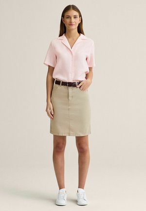 Kvinde står iført en lyserød kortærmet skjorte med knapper, beige nederdel til knæet med brunt bælte og hvide sneakers.