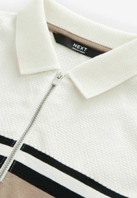 Polo blanc zippé en maille texturée, avec un accent marron au col et des rayures horizontales en noir et beige en bas.