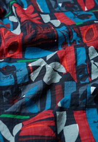 Kolorowa tkanina z geometrycznym wzorem w czerwone, niebieskie, zielone i szare kształty. Materiał ma teksturowany, pognieciony wygląd.