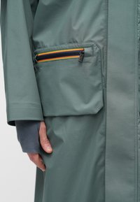 Zelené vonkajšie oblečenie má vrecko na zips s viacfarebným pruhovaným akcentom. Hladká textúra a prispôsobený strih, ktorý zdôrazňuje minimalistický dizajn.