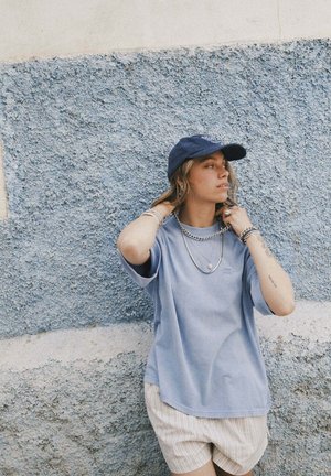 Niebieski bawełniany t-shirt z krótkimi rękawami, noszony z warstwowo nałożonymi srebrnymi naszyjnikami i ciemnoniebieską czapką, zestawiony z jasnymi, paskowanymi szortami.