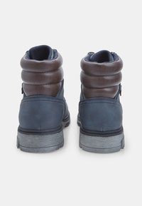 Námořnické modré kotníkové boty s hladkým koženým povrchem, polstrovaným hnědým límcem, texturovanými detaily a robustní šedou podrážkou. Dvě boční zipy.