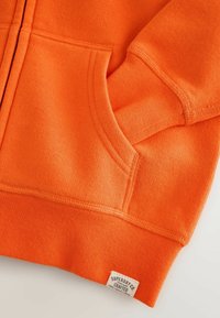 Manche et poche de sweat-shirt orange vif avec bord-côtes et petite étiquette de marque blanche cousue à l'ourlet.