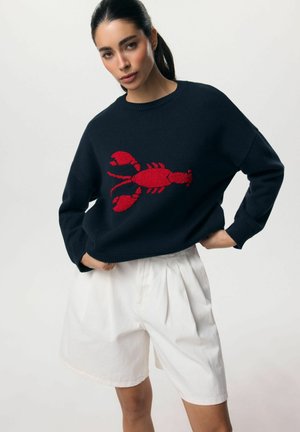 Kobieta z ciemnymi włosami związanymi w kucyk, ubrana w czarny sweter z czerwonym wzorem homara i białe plisowane szorty, pozująca z rękami na biodrach.