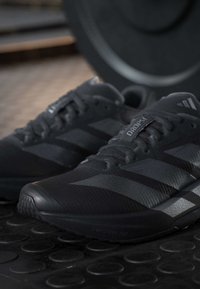 Ζευγάρι μαύρα ελαφριά παπούτσια τρεξίματος Adidas Dropset τοποθετημένα σε υφή γυμναστηρίου με μία βάρος πλάκα στο φόντο.