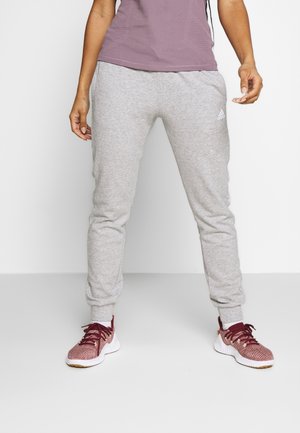 Underkroppen af en person iført lysegrå Adidas joggingbukser, en lilla top, hvide sokker og mørkerøde sports sko, stående mod en ensfarvet baggrund.
