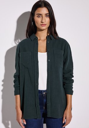 Ciemnozielona koszula z guzikami w teksturowanej tkaninie, z dwoma kieszeniami na piersi i długimi rękawami, noszona na białą bluzkę i zestawiona z niebieskimi dżinsami.