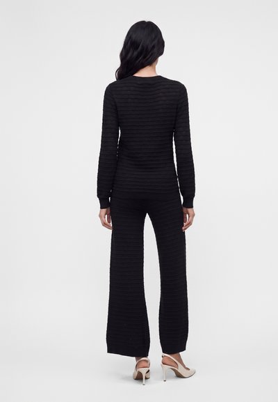 Femme aux longs cheveux foncés portant un haut noir texturé à manches longues et un pantalon à jambes larges, debout de dos, avec des escarpins beige.