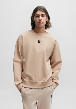 Sweatshirt beige clair avec une coupe décontractée, des poignets et un ourlet côtelés. Présente une petite étiquette en logo noir sur la poitrine. Texture douce.