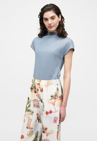 Γυναίκα με σγουρά σκουρόχρωμα μαλλιά, που φοράει ανοιχτό μπλε τοπ με μανίκια σκέλη και λευκό παντελόνι με πολύχρωμα μοτίβα κοκτέιλ και θεμάτων φύσης.