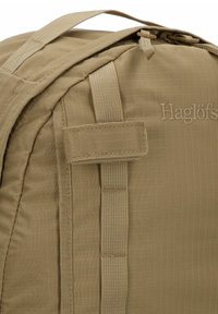Sac à dos khaki avec une surface texturée, doté de sangles renforcées et du logo 'Haglöfs' embouti sur le côté. Design rectangulaire compact.