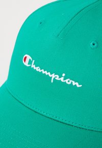 Turkis blå caps laget av teksturert stoff med en buet skygge. Har hvit brodert "Champion"-logo og en liten rød aksent.