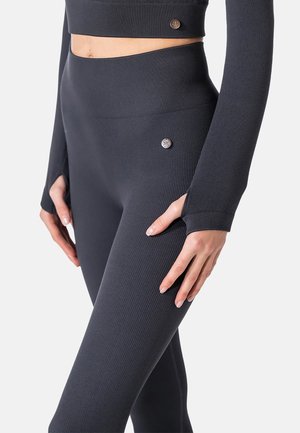 Nahaufnahme einer Person, die dunkle, gerippte Leggings mit hoher Taille und ein passendes langärmeliges Crop-Top mit Daumenlöchern und kleinen metallischen Logo-Pins trägt.