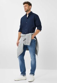 Marineblauw overhemd met knopen en een enkele borstzak, gecombineerd met lichtblauwe spijkerbroek en witte sneakers, afgemaakt met een grijs sweater om de taille gewikkeld.