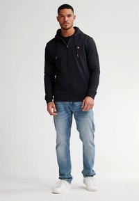 Felpa nera con zip e coulisse, tessuto morbido, vestibilità slim, abbinata a jeans azzurri e sneakers bianche. Design minimalista con accento sul logo.