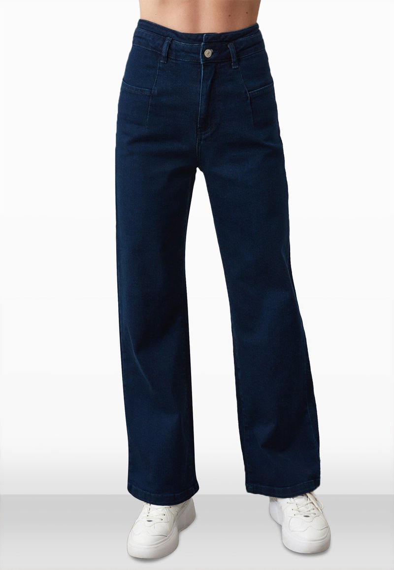 Jean large taille haute bleu foncé porté avec des baskets blanches, visible de la taille aux pieds sur un fond uni.
