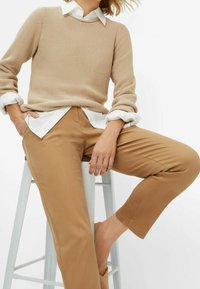 Beige strikket sweater med rund hals, lagdelt over en hvid skjorte. Tan bukser med lige snit og ankellængde, båret uden sko.