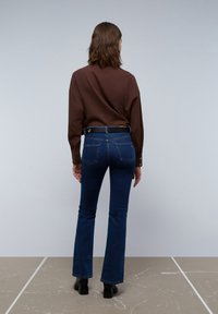 Brun langærmet skjorte, mørkeblå flagrende jeans, set fra ryggen. Jeans med lommer, let falmende, iført sorte ankelstøvler på en neutral gulv.