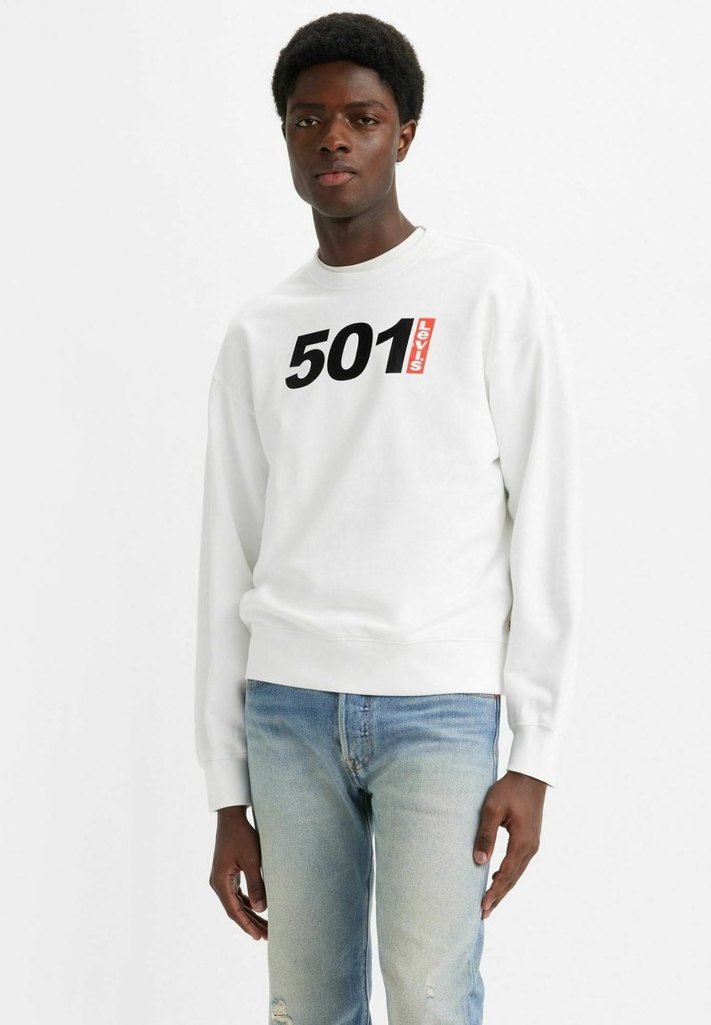 Felpa bianca con grafica nera "501" e logo rosso Levi's. Indossata con jeans chiari strappati, dotati di collo rotondo e maniche lunghe.