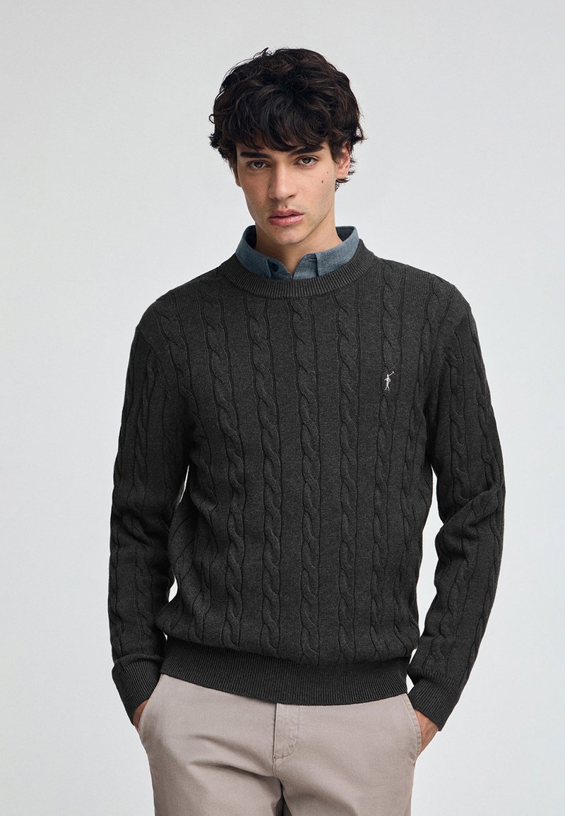 Kabelstrickpullover in Anthrazit mit Rundhalsausschnitt, strukturiertem Muster und schmalen Bündchen. Kombiniert mit einem hellblauen Hemd mit Kragen darunter.