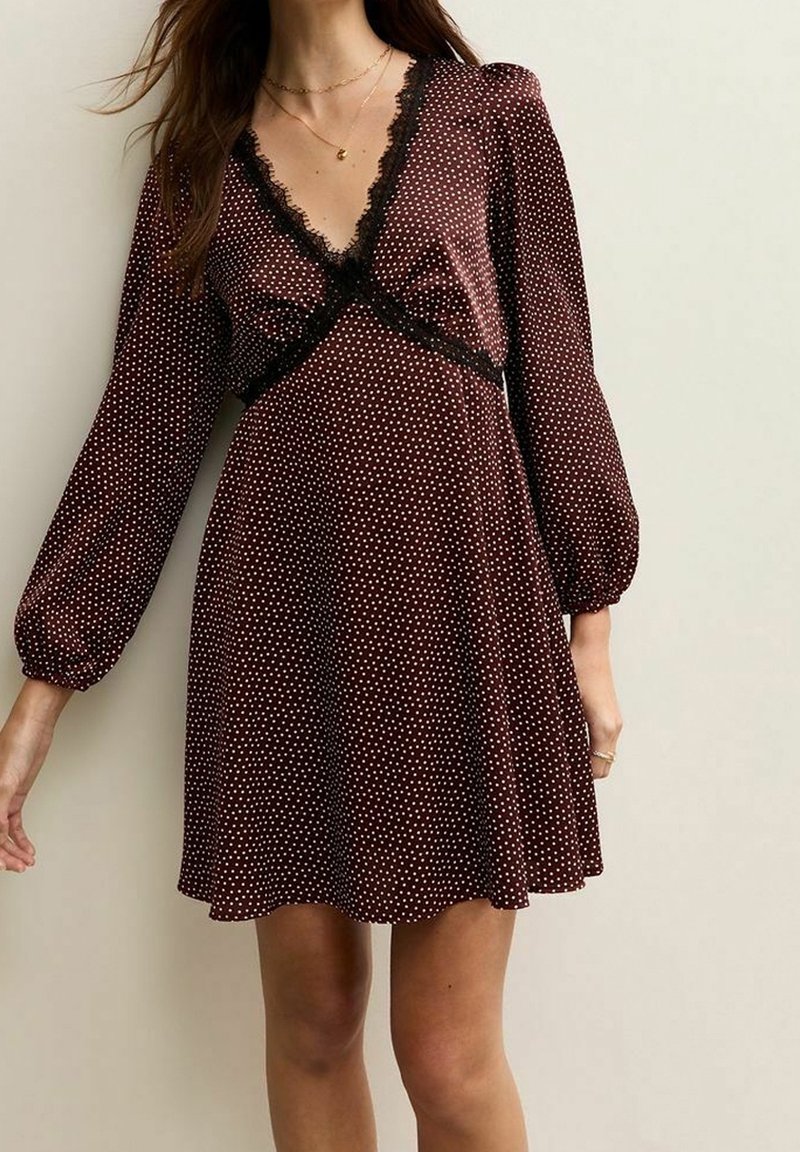 Femme portant une robe marron à pois avec une bordure en dentelle noire, des manches longues bouffantes et un décolleté en V, debout devant un fond clair uni.