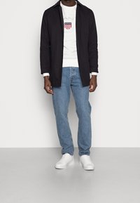 Camisa negra de manga larga con botones, camiseta gráfica blanca, jeans azules y zapatillas blancas, exhibidos contra un fondo neutral.