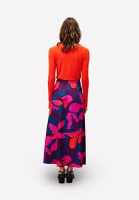 Frau steht mit dem Rücken zur Kamera, trägt ein leuchtend orangefarbenes Langarmoberteil und einen langen Rock mit kräftigem, pink-rot-blauem Blumenmuster sowie schwarze Schuhe.