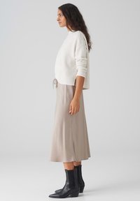 Hellbeige Strickpullover mit lockerer Passform, kombiniert mit einem seidigen beige Midi-Rock und schwarzen Ankle Boots mit spitzem Schuh.