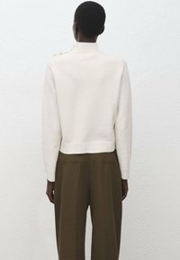 Weißer Strickpullover mit hohem Kragen, gerippten Bündchen und cropped Saum, kombiniert mit olivgrünen, hoch sitzenden Hosen mit Hintertaschen.