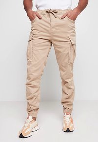 Pantalon cargo beige avec poignets élastiques, plusieurs poches latérales et taille réglable avec cordon de serrage. Le tissu semble lisse avec une finition durable.