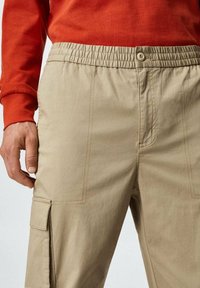 Khaki cargobroek met een elastische tailleband en knoopsluiting, voorzien van zijzakken en zichtbare stikdetails.