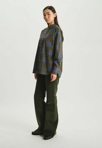 Chemisier à manches longues avec des motifs floraux bleus et verts, associé à un pantalon en velours côtelé vert taille haute. Chaussures simples et coiffure élégante.