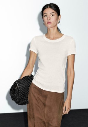 Vrouw die tegen een witte muur staat, gekleed in een witte top met korte mouwen, een bruine rok, zilveren ringoorbellen, en een zwarte geweven clutch tas vasthoudt.