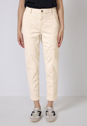 Pantaloni beige chiaro con taglio dritto, dotati di tasche frontali, cuciture e dettaglio con bottone in metallo. Abbinati a sneakers grigie e nere.