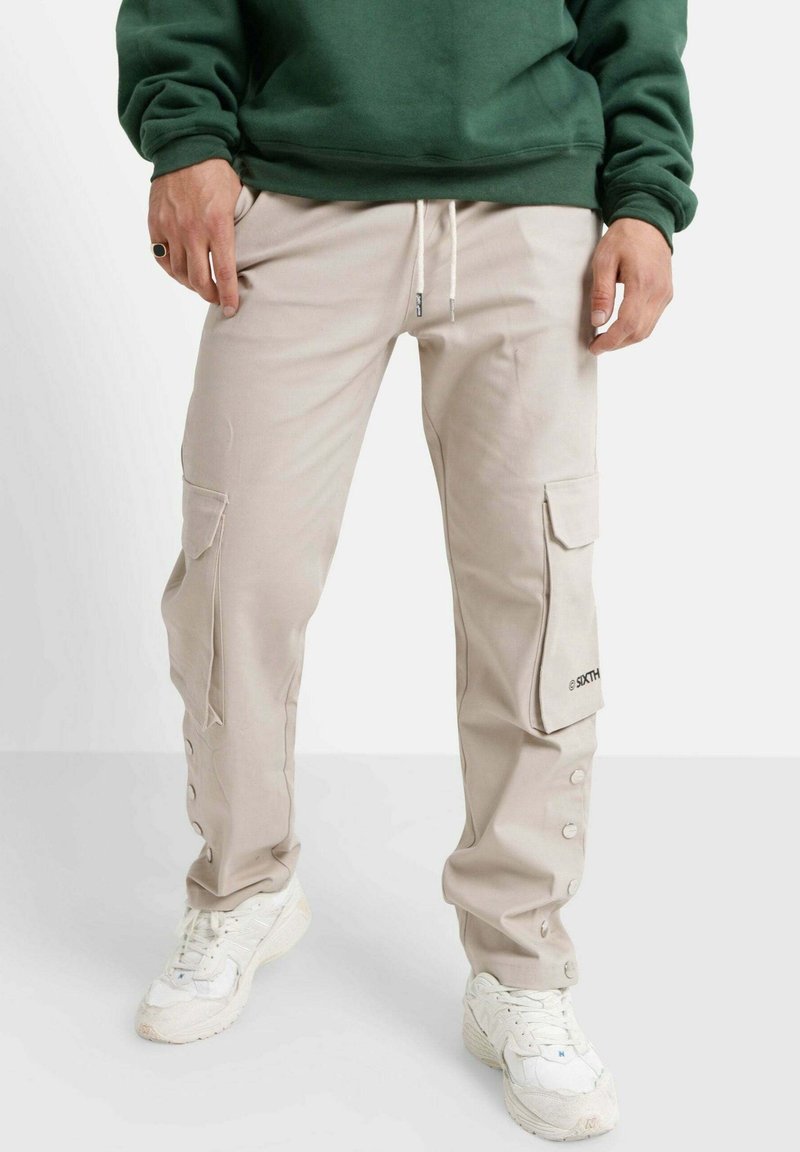 Beige Cargohosen mit Seitentaschen, Kordelzugbund und Druckknöpfen an den Bündchen, kombiniert mit weißen Sneakers und einem grünen Sweatshirt.