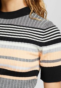 Raidallinen neulepaita, jossa on pystysuuntaista ribbineulosta mustissa, harmaissa, valkoisissa ja persikan värisissä sävyissä. Lyhyet hihat ja musta kaulus.