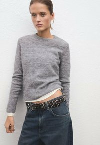 Siva pletena pulover s krožnim izrezom, nošen čez belo srajco. Kombiniran z temno denim hlačami in črnim pasom z srebrnimi kovinami.