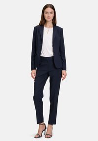 Completo blu navy su misura con giacca aderente e pantaloni dritti, abbinato a una camicia bianca. Presenta un design elegante e accenti minimalisti.