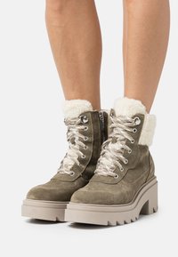 Bottines en suede olive avec un col en fausse fourrure blanche, des lacets argentés et une semelle épaisse beige avec un motif en zigzag.