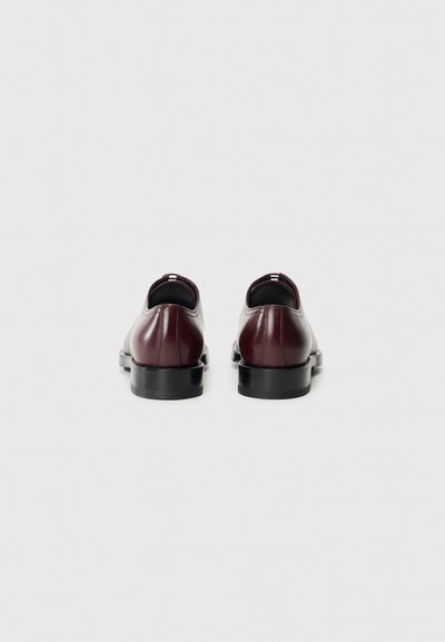 Scarpe in pelle bordeaux con una finitura liscia, forma arrotondata e suole in gomma nera. Presentano un design minimalista senza motivi visibili.