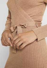 Abito in maglia di un morbido beige con texture a costine. Presenta un design avvolgente e una cintura annodata in vita per una silhouette definita. Maniche lunghe.