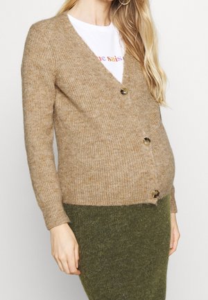 Femme portant un cardigan beige boutonné sur un t-shirt blanc à motif graphique et une jupe duveteuse vert olive, debout devant un fond uni.