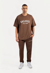 T-shirt en coton marron avec l'inscription "SMILODOX ATHL.WEAR", associé à un jogging assorti. Comprend des baskets blanches, une coupe décontractée, sans motifs visibles.