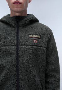Giacca in pile di colore verde scuro con superficie texturizzata, dotata di chiusura con zip, cappuccio e una patch con logo e un accento a bandiera sul petto.