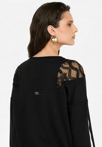 Sweatshirt noir avec des panneaux en dentelle aux épaules et un petit logo doré. Il présente une coupe décontractée et des manches longues avec une texture douce.