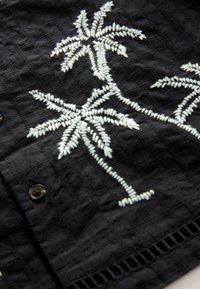 Schwarzer Stoff mit weißen, gestickten Palmen. Die Textur ist leicht erhaben und weist ein kariertes Muster sowie Knopfdetails auf.