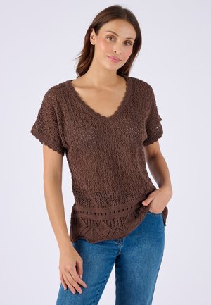 Vrouw die een bruin gebreid topje met korte mouwen en V-hals draagt en een blauw spijkerbroek, staand tegen een effen lichte achtergrond.