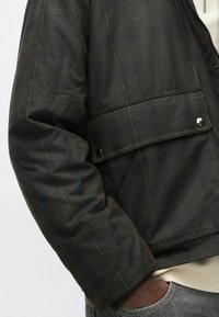 Nahaufnahme einer Person, die eine dunkelgrüne karierte Jacke mit Klappen-Tasche und Druckknöpfen trägt, während die Hand in der Tasche einer grauen Jeans ist.