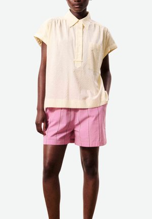 Personne portant un chemisier ample jaune pâle à manches courtes avec un col et un short rose à rayures verticales, debout avec une main dans la poche.