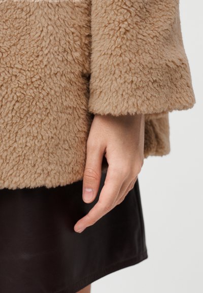 Ταν, αφράτο, υφασμα με υφή ενός ζεστού εξωτερικού ρούχου, με τελείωμα στον καρπό και ένα χέρι που ακουμπάει δίπλα σε μια σκοτεινή καφέ δερμάτινη φούστα.